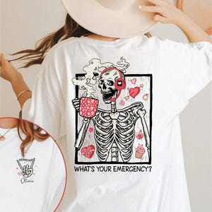 May include: White t-shirt with a graphic of a skeleton wearing headphones and holding a mug of coffee. The skeleton is surrounded by hearts and other Valentine's Day themed imagery. The text "What's your emergency?" is printed below the graphic.