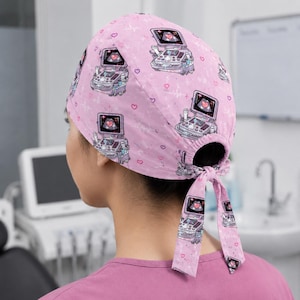 Peut inclure: Une charlotte de bloc rose clair avec un motif répétitif de machines à échographie médicale affichant des icônes de cœur, parsemé de petits cœurs et de lignes d'ECG. Le bonnet est froncé à l'arrière avec deux liens en tissu ajustables.