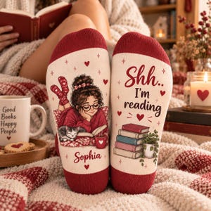 May include: A pair of cream-coloured socks with dark red cuffs and toes. The left sock features an illustration of a person with glasses reading a book with a cat, and the name Sophia. The right sock says Shh.. I'm reading with a stack of books.