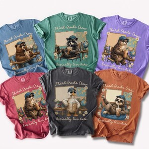 Puede incluir: Seis camisetas de manga corta en azul, verde, morado, rojo, gris oscuro y naranja. Cada camiseta presenta un gráfico de un animal con gorra y gafas de sol, bebiendo de un vaso, con el texto Third Grade Crew y basically live here.