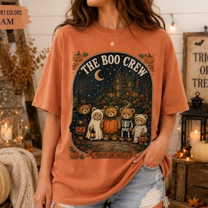 May include: A burnt orange t-shirt featuring a graphic of five teddy bears dressed in Halloween costumes standing in front of a haunted house. The text reads THE BOO CREW and TRICK OR TREAT. The shirt is labelled Comfort Colors Yam.