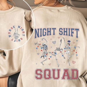 Könnte beinhalten: Beigefarbener Pullover mit dem Text "NIGHT SHIFT SQUAD" in Blau und Rot. Das Design zeigt drei Skelette, die mit rot-weiß-blauen Bändern tanzen. Der linke Ärmel trägt den Text "NURSE NIGHT SHIFT" in Blau.