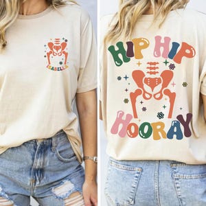 May include: A beige t-shirt with a colourful graphic of a hip bone and the text "Hip Hip Hooray" on the back. The front of the shirt has a smaller version of the graphic with the name "Isabella" underneath.