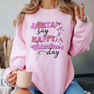 May include: Pink sweatshirt with a graphic of a heart and an aorta with the text "Aorta say happy Valentine's day".