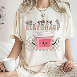 May include: A white t-shirt with a pink cartoon image of a cooler with a smiling face. The text "STAY SHARP" is printed in green and brown above the cooler. The cooler is surrounded by a checkered pattern in green, brown, and white.