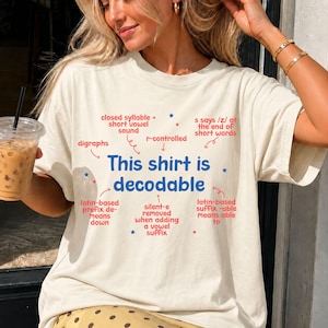 Op de afbeelding: Een crèmekleurig t-shirt met de blauwe tekst "This shirt is decodable" in het midden. Rondom de tekst staan rode pijlen en labels die taalkundige concepten zoals "closed syllable", "digraphs" en "r-controlled" uitleggen met kleine blauwe sterretjes.