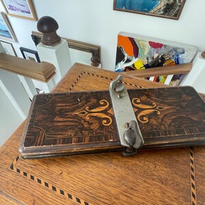 Op de afbeelding: Een vintage houten kist met een decoratief ingelegd patroon in donkerbruine en gouden tinten. Een zilveren metalen sluiting is aan de bovenkant bevestigd. De kist staat op een houten tafel met een vergelijkbaar ingelegd ontwerp.