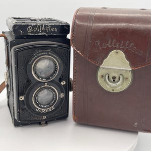 May include: A black Rolleiflex twin-lens reflex camera with a leather strap and a brown leather carrying case featuring the Rolleiflex logo. The camera body displays the text Rolleiflex, Franke & Heidecke, Braunschweig, and Compur on the front.