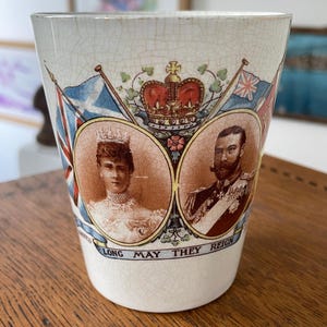 May include: A vintage ceramic cup with a cracked, off-white finish. It features portraits of a royal couple, flags, and a crown. The text "LONG MAY THEY REIGN" is printed on the cup.