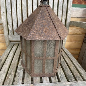 Puede incluir: Una linterna hexagonal vintage con un marco de metal marrón oxidado y un techo puntiagudo. La linterna presenta paneles de vidrio y un lazo de metal para colgar. La linterna está sobre un banco de madera.
