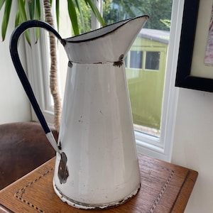 Puede incluir: Jarra de esmalte blanco con asa y borde negros. Esta jarra vintage tiene un pico vertedor y muestra signos de desgaste, lo que le da un encanto antiguo. Se encuentra sobre una mesa de madera.