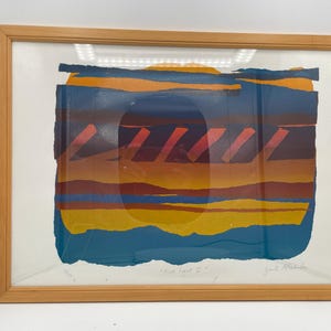 Puede incluir: Una impresión abstracta enmarcada titulada First Light I, que presenta bandas horizontales de azul, naranja y amarillo con trazos rojos diagonales. La obra está en un marco de madera clara con un paspartú blanco y una firma en la esquina.