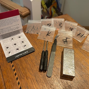 May include: An assortment of lighter flints, tools, and accessories are arranged on a wooden surface. The items include a package of flints, small screwdrivers, tweezers, a metal lighter, and small plastic bags labeled with letters and numbers.