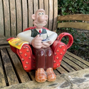 Puede incluir: Una tetera de cerámica con forma de personaje de dibujos animados sentado en un sillón rojo. El personaje sostiene una taza de té y un platillo. La tetera tiene un pico y una manija. El sillón tiene un patrón floral. El personaje lleva una camisa blanca, un chaleco verde, pantalones marrones y zapatos a cuadros. La tetera está sentada sobre una superficie de madera. La tetera tiene una revista en el reposabrazos que dice "British Cheese & Biscuits".