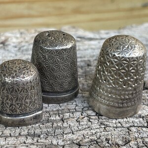 May include: Three silver thimbles with a starburst pattern. The thimbles are sitting on a piece of weathered wood.