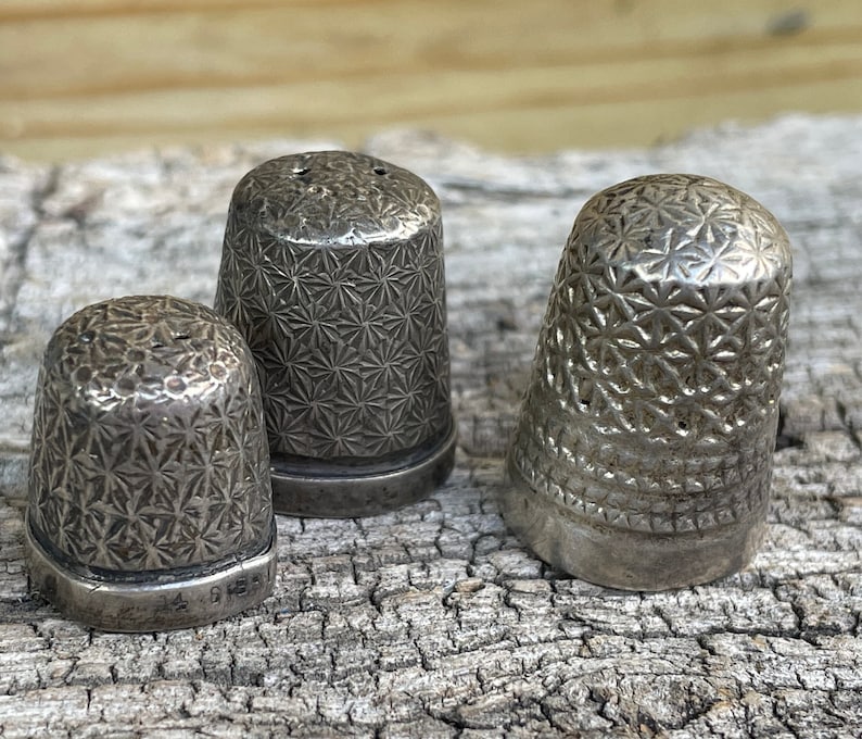 May include: Three silver thimbles with a starburst pattern. The thimbles are sitting on a piece of weathered wood.