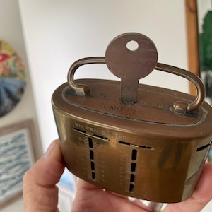Peut inclure: Une tirelire vintage en laiton avec une clé et une poignée. La tirelire comporte des fentes pour les pièces et le texte "HOME SAFE" est visible. La clé est insérée en haut. La tirelire est tenue dans une main.