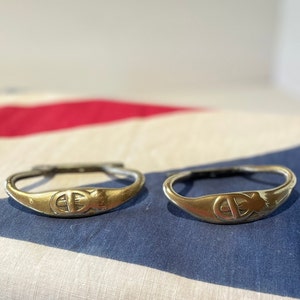 May include: Two gold-colored metal rings with a decorative design on the front. The rings are on a white and blue fabric background.