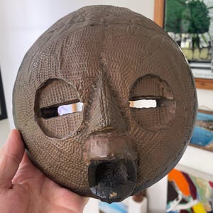 May include: A brown, textured mask with a carved face, featuring eye and mouth openings. The mask has a rounded shape and is held in a hand. The mask's surface has a pattern of small, raised dots. The mask is a decorative item.