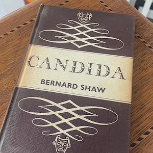 May include: A hardcover book titled "Candida" by Bernard Shaw. The book has a dark brown cover with gold lettering and decorative flourishes. The book is resting on a wooden surface with a patterned design.