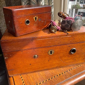 Puede incluir: Dos cajas de madera antiguas con herrajes de latón, una encima de la otra. La caja superior es más pequeña, con una cerradura y una abertura circular. La caja inferior tiene una cerradura y una abertura circular. Varios accesorios antiguos encima.