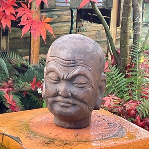 Peut inclure: Une sculpture de tête en bois sculpté représentant un homme avec une expression sérieuse. La sculpture est de couleur brune et a un aspect vieilli. Elle est posée sur un piédestal carré de couleur brun orangé.