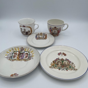 May include: Two ceramic cups and two saucers featuring commemorative royal imagery. The items display portraits of monarchs, British flags, crowns, and text including Silver Jubilee 1910-1935. The pieces are off-white with gold or blue trim accents.