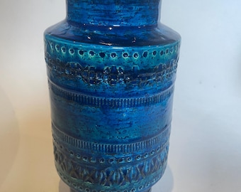May include: A tall, blue ceramic vase with a textured surface and decorative bands. The vase features a gradient of blue hues, from deep teal to bright turquoise. The design includes embossed patterns and a wide base.