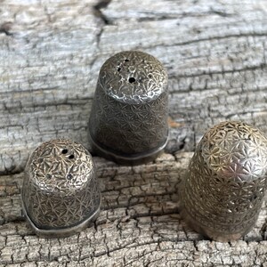 May include: Three silver thimbles with a starburst pattern. The thimbles are arranged in a triangle shape on a wooden surface.
