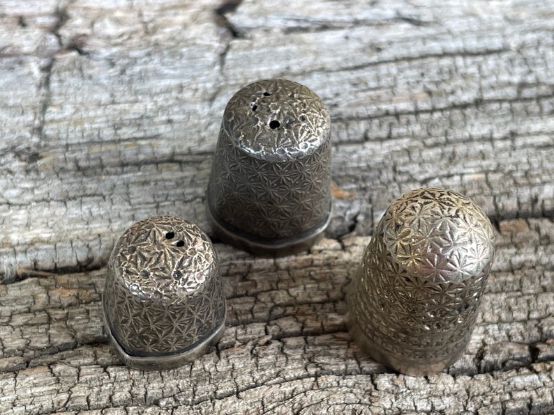 May include: Three silver thimbles with a starburst pattern. The thimbles are arranged in a triangle shape on a wooden surface.
