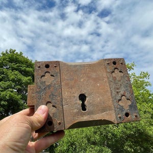 May include: An aged door lock, featuring a keyhole and decorative metal plates. The lock is constructed from wood and metal, displaying signs of wear and rust. The metal plates have a quatrefoil design. The background shows a blue sky and green foliage.