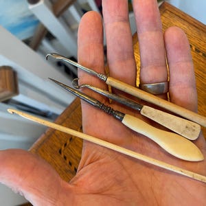 Puede incluir: Una colección de ganchillos y herramientas de ganchillo vintage. Las herramientas tienen mangos de madera y color marfil con extremos de gancho de metal. Las herramientas son de varios tamaños y formas, lo que sugiere diferentes técnicas de ganchillo.