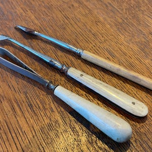 May include: Three antique serving utensils with metal blades and handles. The handles are made of bone or ivory, and the blades are silver-toned. The utensils are arranged on a wooden surface.