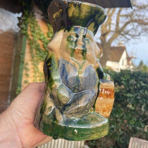 Puede incluir: Una figurita de cerámica de un gnomo con un sombrero verde y marrón, sosteniendo una jarra marrón. El gnomo está sentado sobre una base verde y tiene un esmalte verde, marrón y azul.