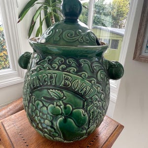 May include: A green ceramic punch bowl with a matching lid. The bowl features embossed fruit designs and the words "PUNCH BOWL". The bowl has two side handles and a gold rim. The lid has a round handle.