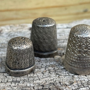May include: Three silver thimbles with a starburst pattern. The thimbles are different sizes and are sitting on a piece of weathered wood.