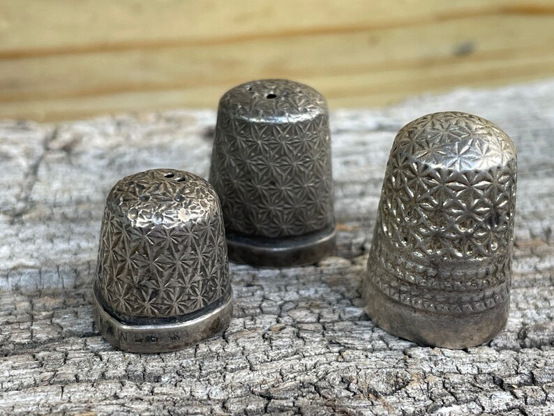 May include: Three silver thimbles with a starburst pattern. The thimbles are different sizes and are sitting on a piece of weathered wood.