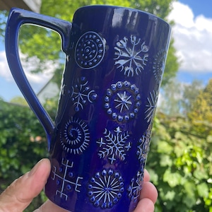 May include: A cobalt blue ceramic mug with a large handle. The mug is decorated with raised white snowflake and circular patterns. The mug is held in a hand against a background of green foliage and a blue sky.
