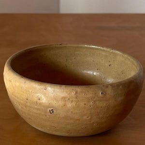 Puede incluir: Un cuenco pequeño y redondo de cerámica con un exterior marrón claro y moteado. El cuenco tiene un interior liso y un borde ligeramente irregular. El color es un tono cálido y terroso, lo que sugiere una estética artesanal o rústica.