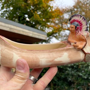 May include: A ceramic canoe planter featuring a Native American figure in a feathered headdress paddling the canoe. The planter is painted in shades of brown and white.