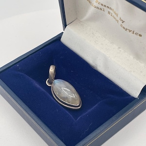 May include: A marquise-shaped moonstone pendant set in a silver-toned metal frame, resting inside a dark blue velvet-lined presentation box. The interior lid of the box features the gold-colored text: Presented by National Blood Service.