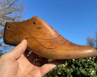 May include: A vintage wooden shoe last, showcasing a classic shoe form. The last is a warm brown color, with visible grain and markings. It has a curved shape, with a defined heel and toe. The shoe last is held against a blue sky background.