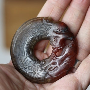 以下が含まれることがあります： リング型の濃い茶色の翡翠のペンダントで、龍のデザインが施されています。龍は細部まで彫られており、表面はテクスチャ加工されています。