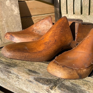Puede incluir: Tres hormas de zapatos de madera, utilizadas por los zapateros para dar forma a los zapatos, están dispuestas sobre una superficie de madera. Las hormas son de madera marrón y tienen un aspecto desgastado y envejecido.
