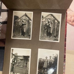 Könnte beinhalten: Ein offenes Vintage-Fotoalbum mit vier Schwarz-Weiß-Fotografien. Die Fotos zeigen Menschen, die im Freien posieren, einige mit Hunden, vor Holzstrukturen. Die Albumseiten sind braun, mit einem rot genähten Rücken.