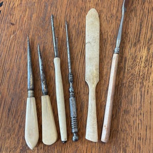 Peut inclure: Ensemble d'outils anciens avec manches en ivoire et lames en métal, comprenant des poinçons, une spatule et une lame incurvée. Les outils sont disposés sur une surface en bois, mettant en valeur leur aspect vieilli et leur savoir-faire.