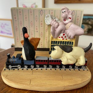 May include: A whimsical figurine featuring a train with a black and white penguin, a yellow dog, and a pink character. The train cars are red and gray. The background has a patterned wallpaper.