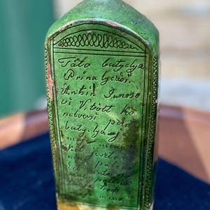 May include: A green ceramic bottle with a rectangular shape and a rounded top. The bottle has a label with handwritten text in black ink. The base of the bottle is yellow and the top is brown.