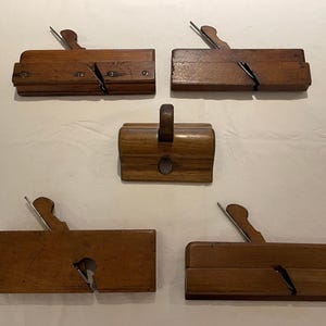 May include: Five antique wooden hand planes. The planes are made of dark wood and have metal blades. The planes are arranged on a light-coloured surface. The planes are used for woodworking and carpentry.