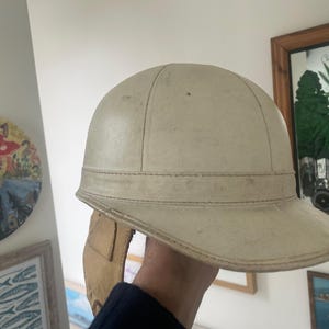 May include: A cream-coloured vintage helmet with a brim and a band around the crown. The helmet appears to be made of leather or a similar material, with visible stitching. It has a classic, old-fashioned design, suggesting it may be a riding or sports helmet.